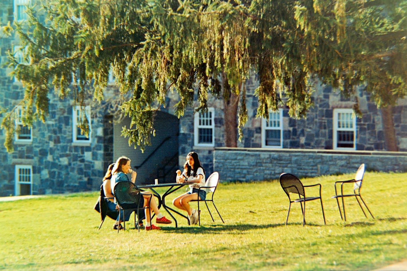 Two friends talking at an outdoor table next to a stone campus building